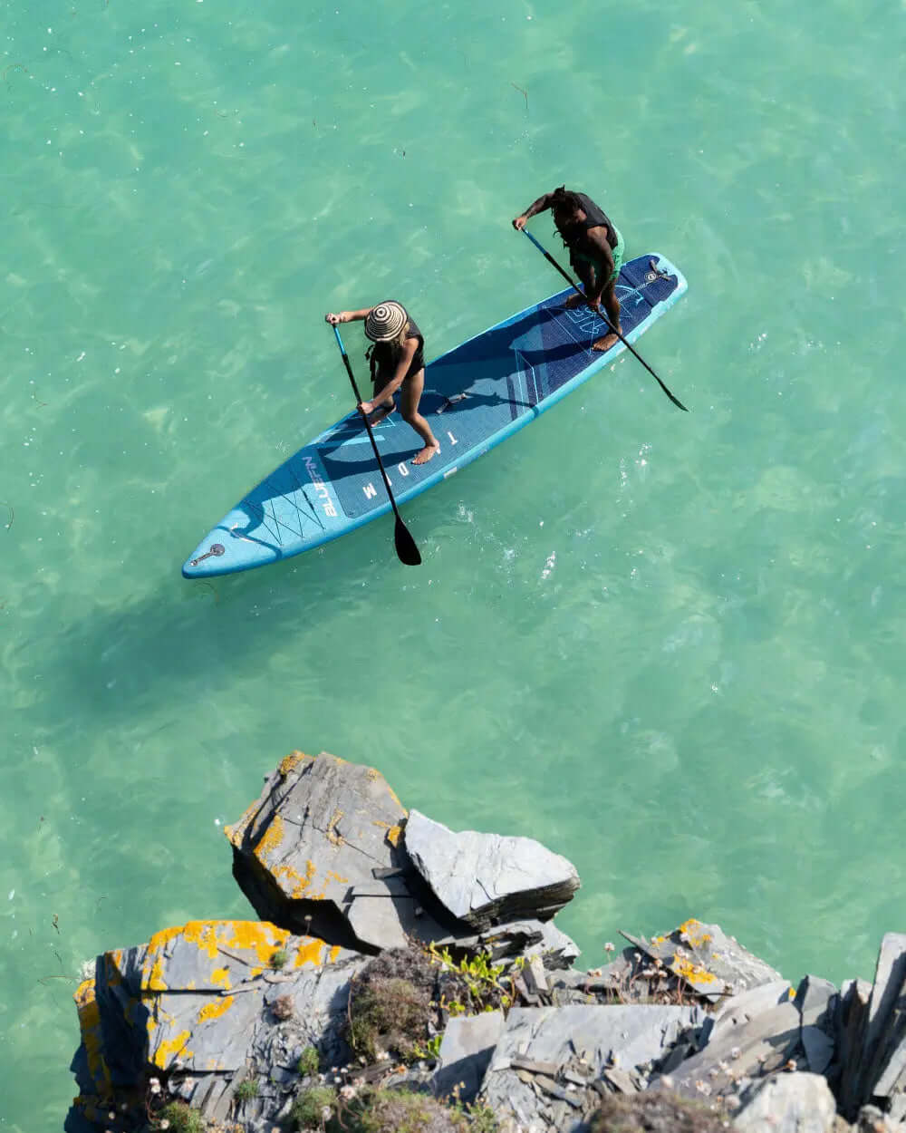Two people paddling on a blue stand-up paddleboard in clear water near rocky shore