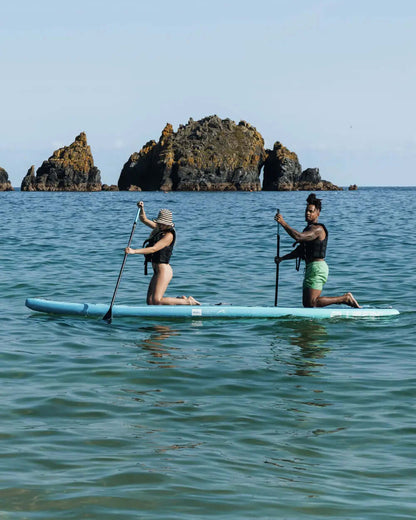 Two people paddleboarding on a blue inflatable board in clear water near rocky islands.