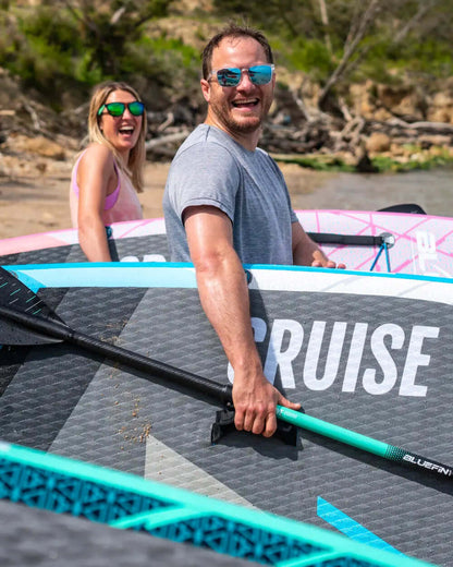 Two people smiling by inflatable paddleboards on a beach, one holding a paddle with a turquoise shaft.